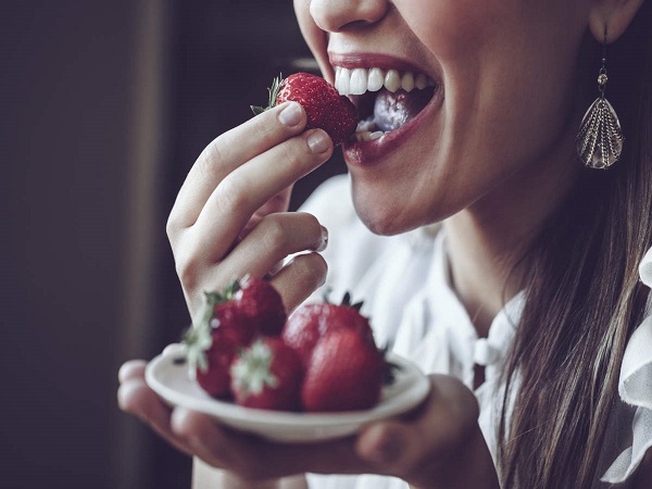 A young women holding a small plate with strawberries and biting a strtwaberry with her white teeth.